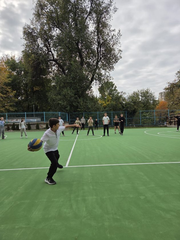 Момче в бяла тениска замахва с волейболна топка на зелено игрище, зад него стоят други ученици